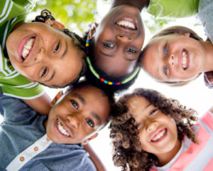 group of children forming a circle with their heads, leaning forward and looking down at the camera. They are all smiling.