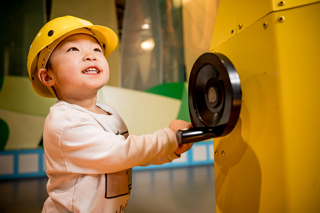 Toddler wearing a yellow construction hat and turning a black crank